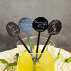 Personalized cocktail stirrers with names and dates in a glass of yellow liquid on a textured surface.