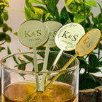 Gold and silver drink stirrers with 'K&S' initials and dates in a glass of liquid, surrounded by greenery.