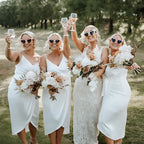 Four women in white dresses holding bouquets and glasses outdoors.