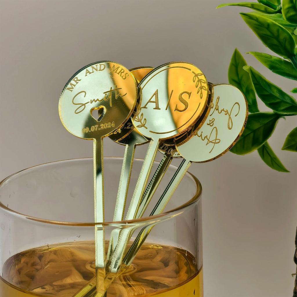 Gold drink stirrers with personalized names in a glass of yellow liquid.