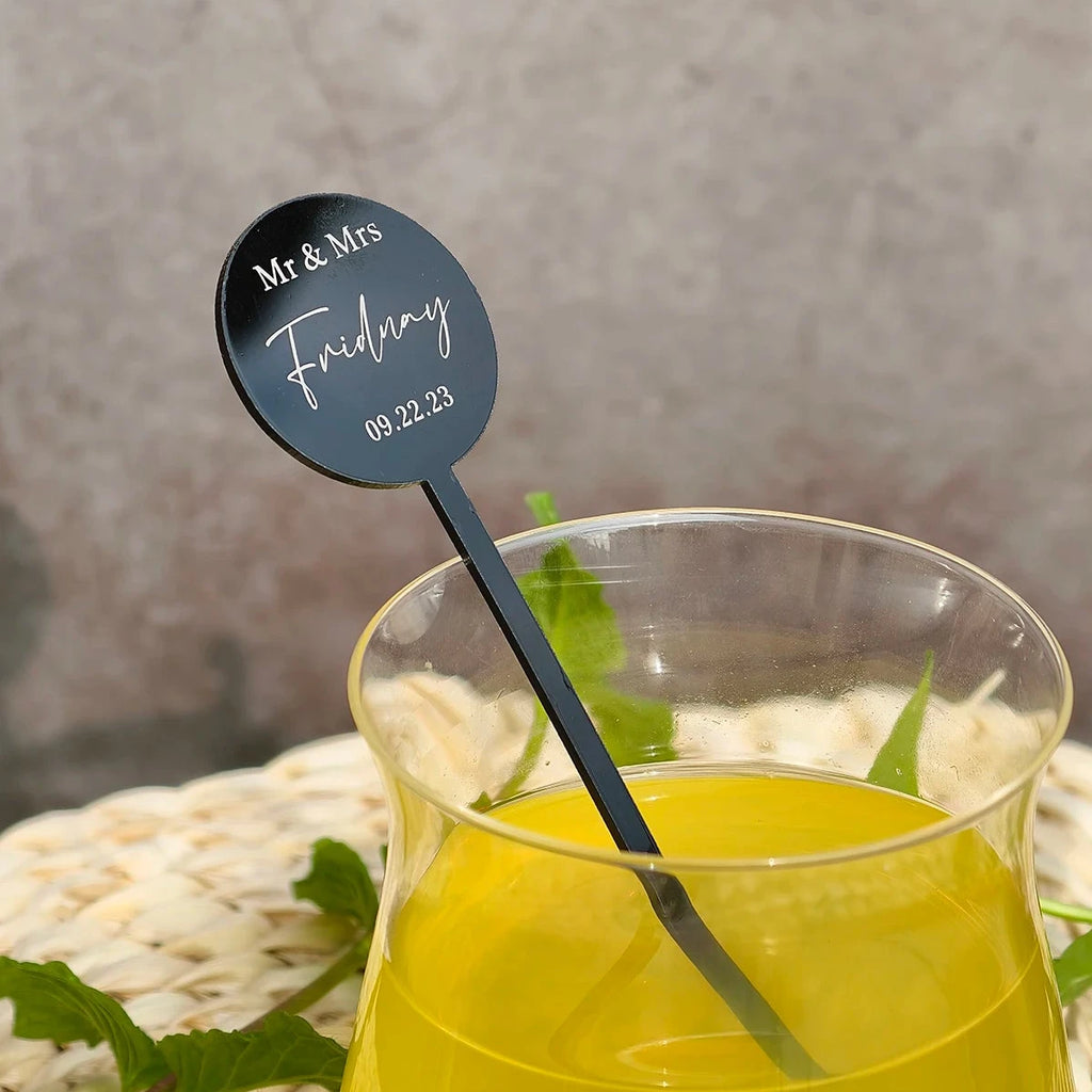 Glass of yellow liquid with a black stirrer labeled 'Mr & Mrs' on a textured surface.