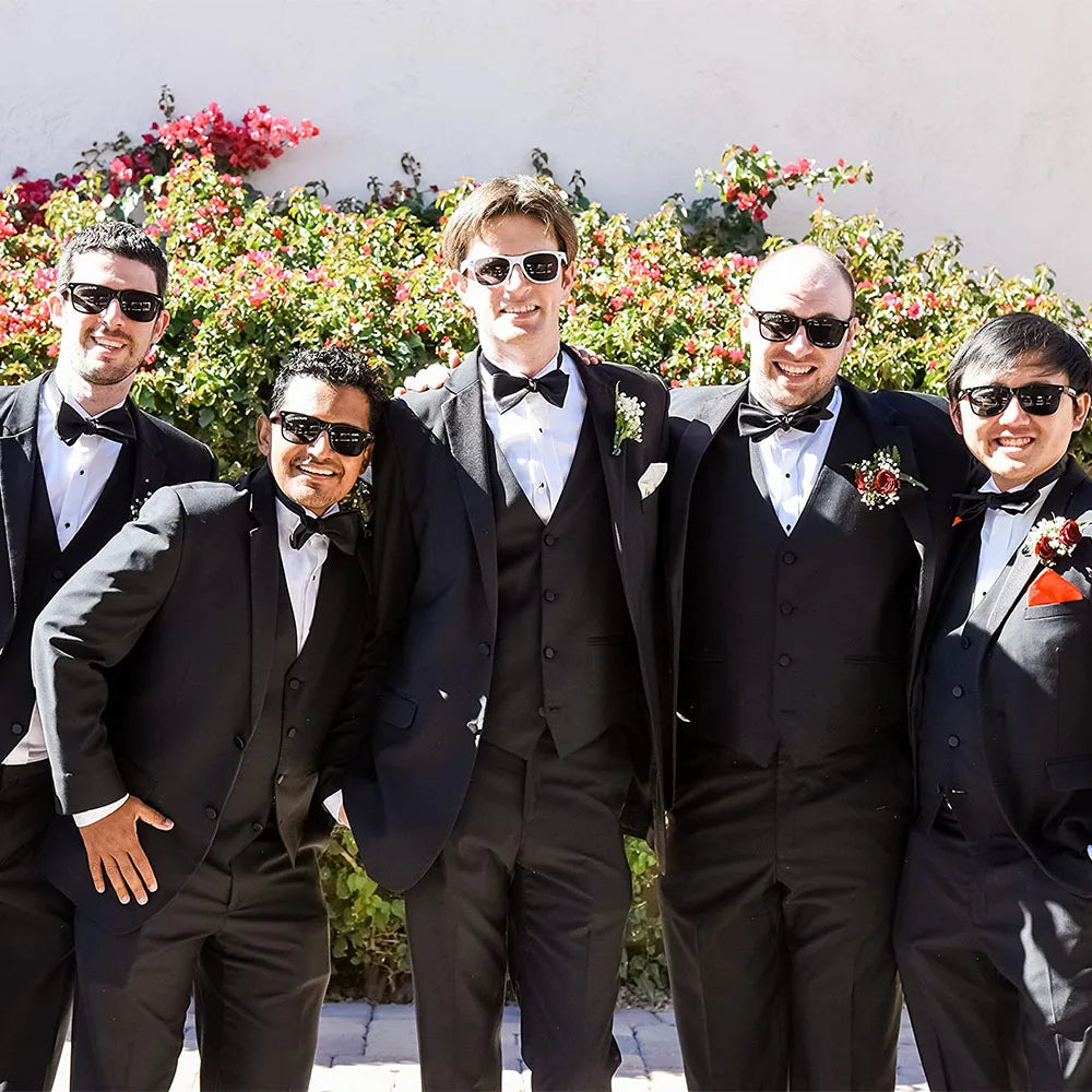 Five men in formal black suits with bow ties standing outdoors against a floral background.