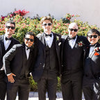 Five men in formal black suits with bow ties standing outdoors against a floral background.